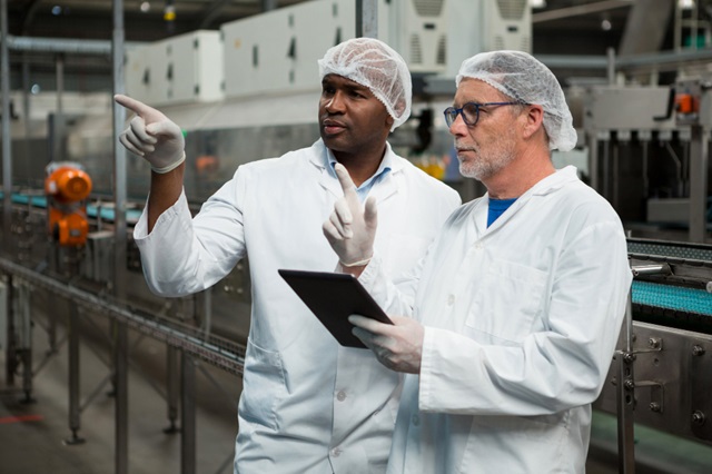 45,000 UK Seasonal Workers Visa Sponsorship Companies and Agencies (Sponsors) 2024/2025 to 2029 for Nigerians and Other Asian Countries including Nepalese, Indonesians, Indians, Pakistanis, and Bangladeshi Images: 1. Two workers, an African-American man and a white man, both wearing white lab coats, hair nets, and gloves, inspecting a food processing production line. The African-American man is pointing while holding a digital tablet.